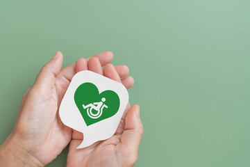 A real white speech bubble paper cut with disabled people inside heart icon on hands, green background including copy space