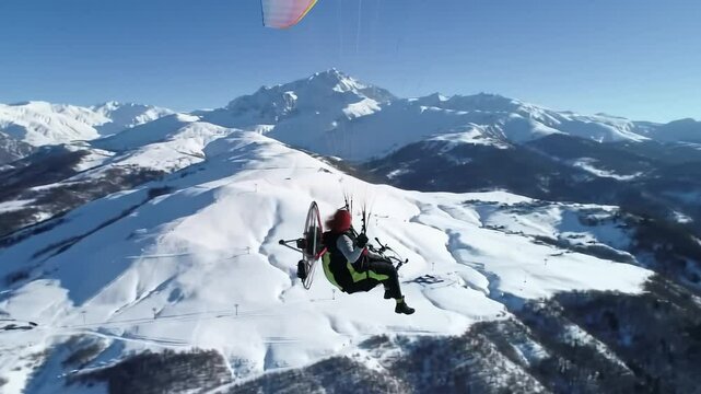 Soaring through the chilly winter air, a paramotor pilot glides above stunning snow-covered peaks. The mountains stretch endlessly under a bright blue sky, showcasing a perfect day for adventure.