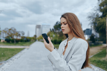 Businesswoman in a gray blazer checking her phone outdoors, with a focused expression and a modern cityscape backdrop.