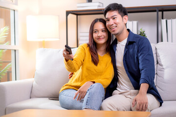 Obraz premium Happy young asian couple sitting on cozy sofa watching TV together and enjoying a relaxation in living room at home, relationship and comfortable of couple during watching television on couch.