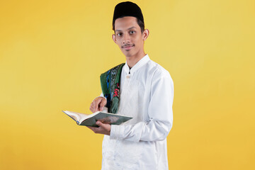 Muslim man wear white shirts with Islamic ornaments and a prayer mat on the shoulder while holding and pointing to Holy book of Quran