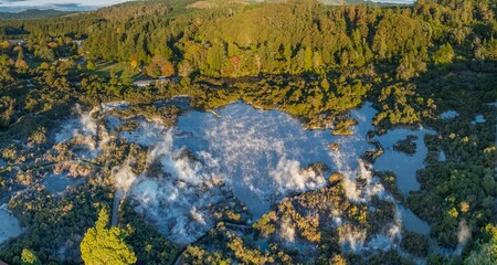Aerial view of Wai-O-Tapu geothermal area, near Rotorua, New Zealand. Steam rises from the hot springs, surrounded by lush forest. Tourists visit to experience the unique landscape.