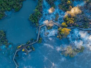 Aerial view of the Wai-O-Tapu geothermal area in New Zealand. The wooden boardwalk allows tourists to safely view the hot springs and mud pools. Steam rises from the geothermal activity.