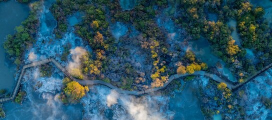 Aerial view of a wooden boardwalk winding through the geothermal landscape of Wai-O-Tapu in New Zealand. Steam rises from the hot springs, attracting tourists.