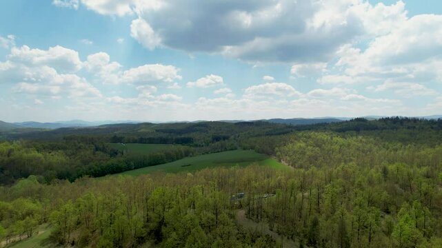 Aerial view of a vast green forest with open fields and rolling hills extending into the distance under a partly cloudy sky, showcasing natural beauty, tranquility, and diverse landscapes