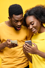 Fototapeta premium Young African couple joyfully interacting with smartphones against a bright yellow background, showcasing excitement and modern lifestyle choices