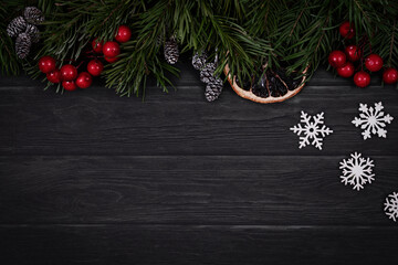 Christmas tradition flat lay with copy space. Atmospheric upper border with green spruce, orange slice, cones, white snowflakes and holly berries on black wooden background.