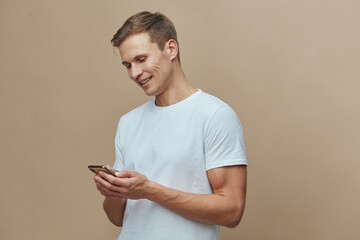 Smiling young man in casual white t-shirt using mobile phone, cheerful expression, engaging with device, casual indoor setting, beige background, lifestyle, everyday life, modern technology concept
