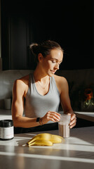 A white woman in her 30s prepares a protein shake in a stylish kitchen, with bananas and a supplement jar on the counter, showcasing a healthy lifestyle and focus on fitness