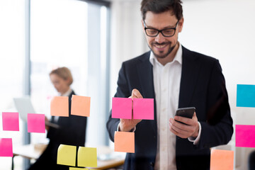 Adult male organizing tasks with colorful sticky notes on glass board, holding smartphone. Office backdrop with modern workplace vibes.