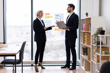 Business meeting scene with adult man and woman discussing project in bright office. Features teamwork, collaboration, professional attire, modern workplace with whiteboard, bookshelves © sofiko14