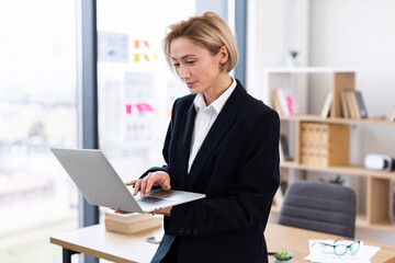 Confident adult woman in formal suit using laptop in bright modern office workplace, focusing on work tasks. Represents business, productivity, technology, and professional career development.