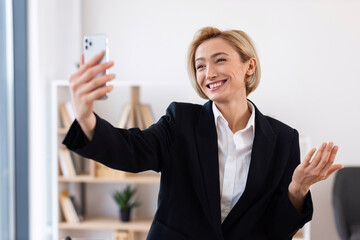 Smiling businesswoman in formal black suit conducting remote video call in bright office...
