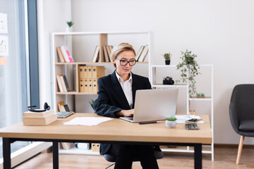 Businesswoman wearing black suit sitting at desk using laptop in bright office setting. Office...