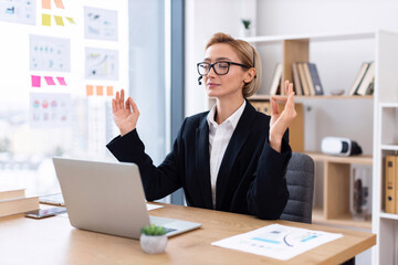 Blond businesswoman wearing glasses meditating at desk in office workplace during work break, promoting relaxation and mindfulness during busy professional environment.