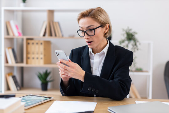 Professional adult Caucasian woman wearing formal suit sitting at desk expressing surprise while looking at smartphone screen in modern office with organized shelves in background.