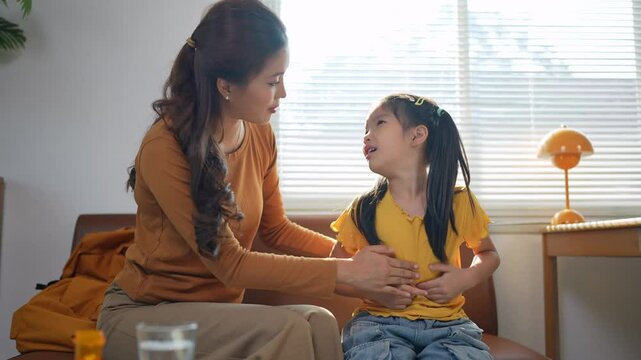 Caring mother gently soothing her preteen daughter experiencing abdominal pain, attempting to administer liquid medication while child resists taking prescribed treatment at home