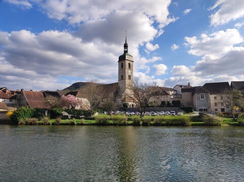 A picturesque riverside town is dominated by a tall church tower with a clock face, surrounded by charming houses and lush greenery. Fluffy clouds float in a bright blue sky, peaceful rural setting.
