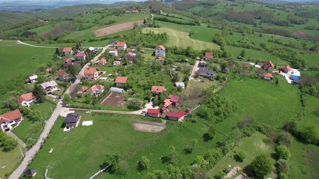 Peaceful rural village surrounded by lush green fields, rolling hills, and scattered red-roofed houses, connected by narrow roads and pathways amidst nature