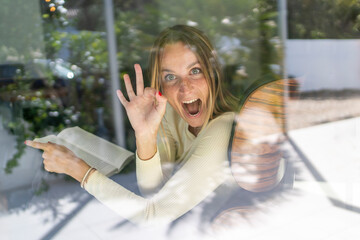 Happy playfull woman by the window at home, doing OK gesture and smiling with a book on her hands