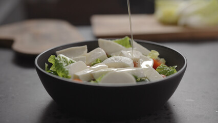 pour walnut oil onto salad with romaine lettuce, tomatoes and mozzarella in black bowl on concrete countertop
