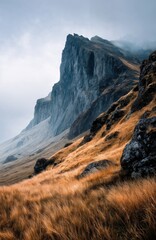 Misty Cliffside Mountain Rising Above Golden Grass Hillside in Autumn Light, Dramatic Landscape with Steep Rocky Slopes, Rugged Nature Scene Evoking Solitude, Wilderness, and Majestic Elevation