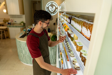 Young storekeeper arranging fresh vegan food inside refrigerated display in his health food store