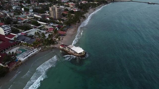 Drone orbiting over Pampatar&rsquo;s coastal town revealing a rocky pier restaurant and beachfront villas lining white sand shore under clear blue sky above turquoise Caribbean waters at midday real time