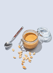 Peanut butter in glass jar on blue background with spoon and salted nuts. Concept of tasty and healthy snack