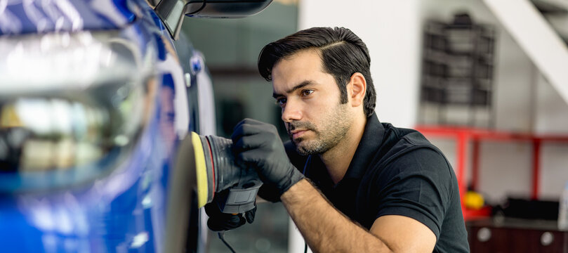 Professional car detailer wearing black gloves and a black polo shirt polishes a blue vehicle using a power buffer. The background features a modern garage with white stairs and equipment