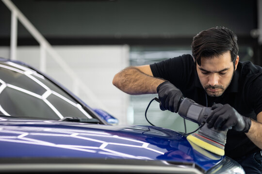Professional car detailer wearing black gloves and a black polo shirt polishes a blue vehicle using a power buffer. The background features a modern garage with white stairs and equipment