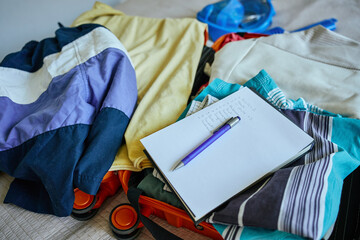 Handwritten checklist and pen on top of colorful folded clothes inside open suitcase. Concept of travel planning and packing essentials for trip