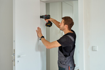 Man holds cordless drill and installs white door handle in modern interior. Handyman fitting door...