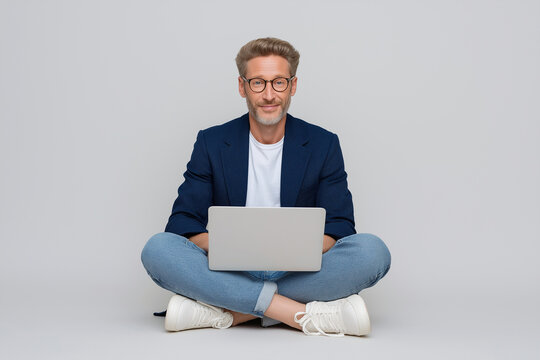 Photo of a mature, handsome man 50 years old sitting on the floor with a laptop, wearing a blue blazer, jeans, and white sneakers. He is also wearing glasses and is isolated against a light gre