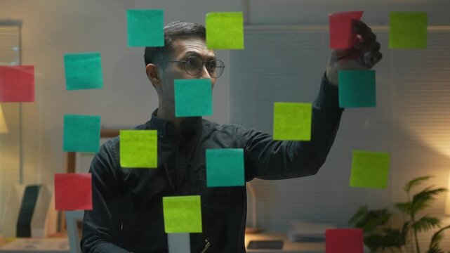A man is standing in front of a window with a green and yellow sticker on it. The stickers are arranged in a pattern and the man is looking at them. Concept of creativity and organization