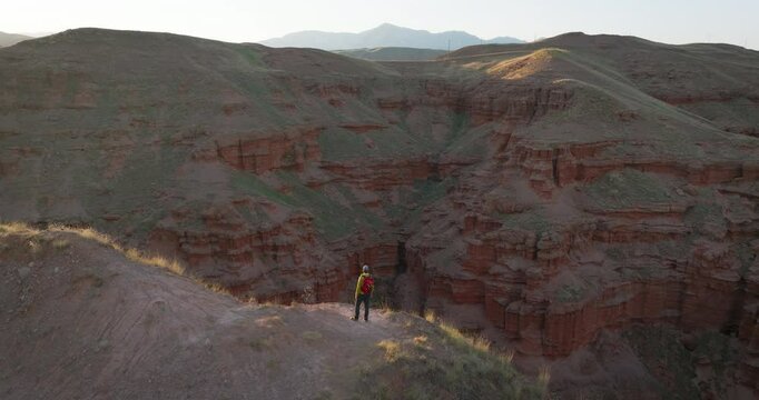 A backpacker observes impressive geological formations among red fairy chimneys in the Narman district of Erzurum. A drone circles him at sunset, recording the moment from above.