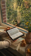 Person reading an open Bible by the window at sunset, surrounded by cozy textures, a candle, yellow...