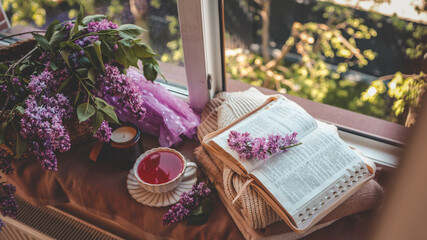Cozy spring still life with lilac branches, an open Bible, a cup of herbal tea, and a candle on a windowsill, bathed in soft natural light and surrounded by warm textures and purple tones