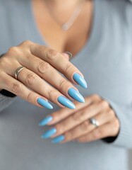 A close-up of a woman's hands featuring long, almond-shaped nails painted in a soft light blue polish, adorned with a silver ring on one finger, against a blurred background.