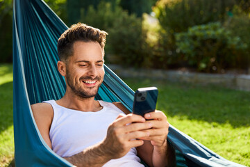 Handsome man using phone in hammock in backyard garden
