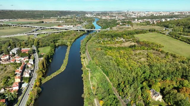 Drone shot - a river winds through a beautiful green landscape - a bridge stretches across the river, and farther in the background a cityscape