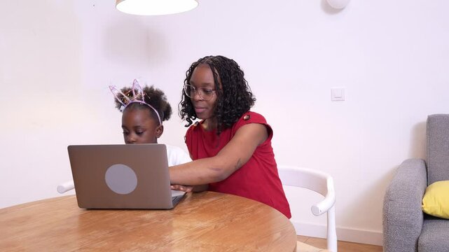 Young african mother assisting her daughter with a laptop, fostering early digital literacy in a comfortable home setting