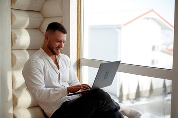 A man is working on a laptop by a large window. Man sitting in chair with laptop and smiling while working by a large window on a sunny day.