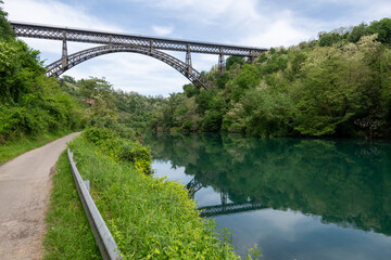 Obraz premium San Michele Bridge spans the Adda River with its elegant iron arch, one of the world’s tallest single-span iron bridges, standing proudly amid lush hills and reflective waters.
