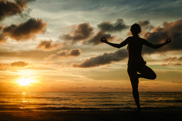 Fototapeta premium A woman practicing yoga while standing on a tropical beach during a stunning sunset, surrounded by serene beauty and warm light.