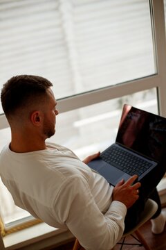 A man is working on a laptop by a large window. Man sitting in chair with laptop and smiling while working by a large window on a sunny day.
