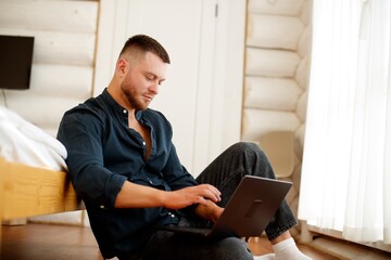 A man with a laptop in a cozy house. A man sits on the floor, focused on his laptop, surrounded by a cozy home environment with wooden walls.