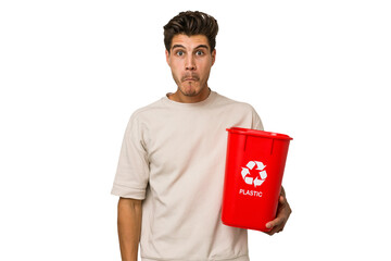 Young caucasian man holding a plastic trash isolated shrugs shoulders and open eyes confused.