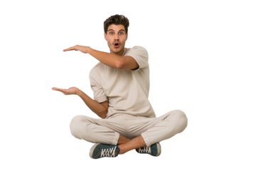 Young caucasian man sitting on the floor isolated on white background shocked and amazed holding a copy space between hands.