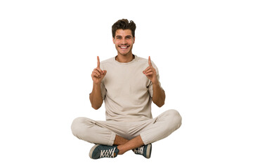 Young caucasian man sitting on the floor isolated on white background indicates with both fore...